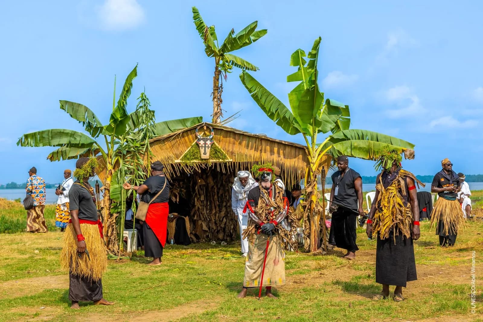 A group of people in traditional dress and raffia costumes outside a thatched hut among banana trees.